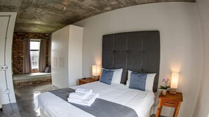 A modern bedroom with a large bed, a headboard with a striped pattern, bedside lamps, and a small table with a plant. - Villa Sunset Mansion Cape Town (Cape Town)