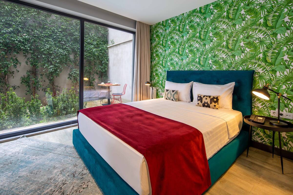 A hotel room with a large bed, a view of greenery through a window, a small table with a chair, and a wall with a leafy pattern.