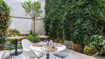 Un área de comedor al aire libre con mesas redondas, sillas blancas y un muro cubierto de vegetación.