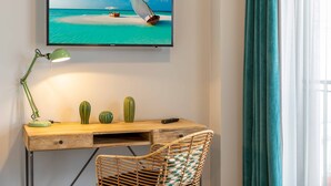 A room with a wooden desk, a wicker chair, a television displaying a beach scene, a green desk lamp, and a remote control. - Green Parrot Hotel (Istanbul)