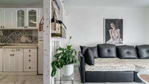 Une cuisine moderne avec des meubles blancs et un pare-feu en carrelage, un canapé en cuir noir agrémenté d’une housse en forme de journal et un tableau encadré au mur.