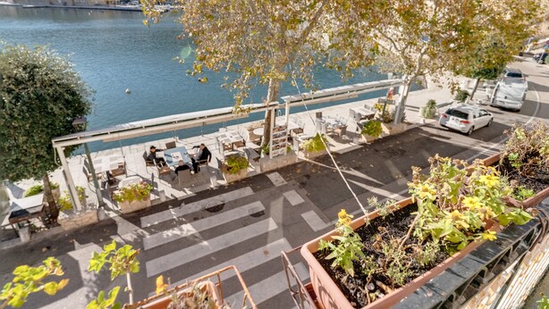Une terrasse avec des sièges extérieurs donnant sur une étendue d’eau, entourée de plantes en pot et offrant une vue sur des bâtiments au loin.