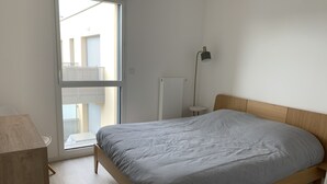 A bedroom with a wooden bed, a bedside table, a lamp, and a window with blinds. - Cozy 1BR Apartment in Central Fort-Mahon-Plage with Private Parking (Fort-Mahon-Plage)