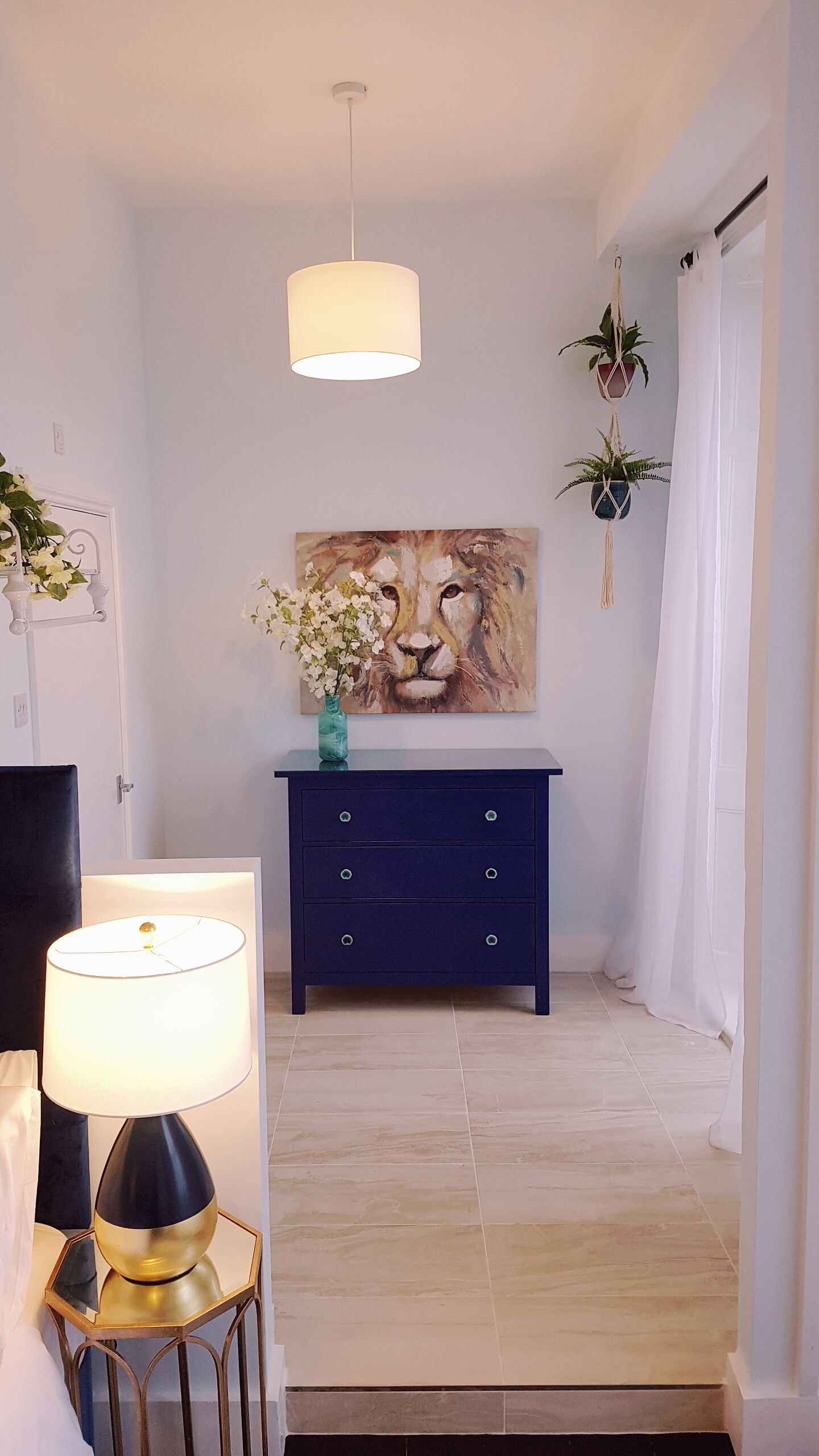 Un couloir avec une commode bleue, un tableau représentant le visage d’un lion et une plante suspendue.