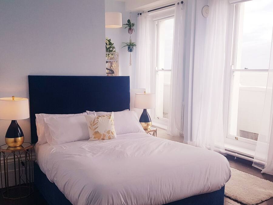 A bedroom with a large bed, two bedside tables, a wall-mounted lamp, and a window with sheer curtains.