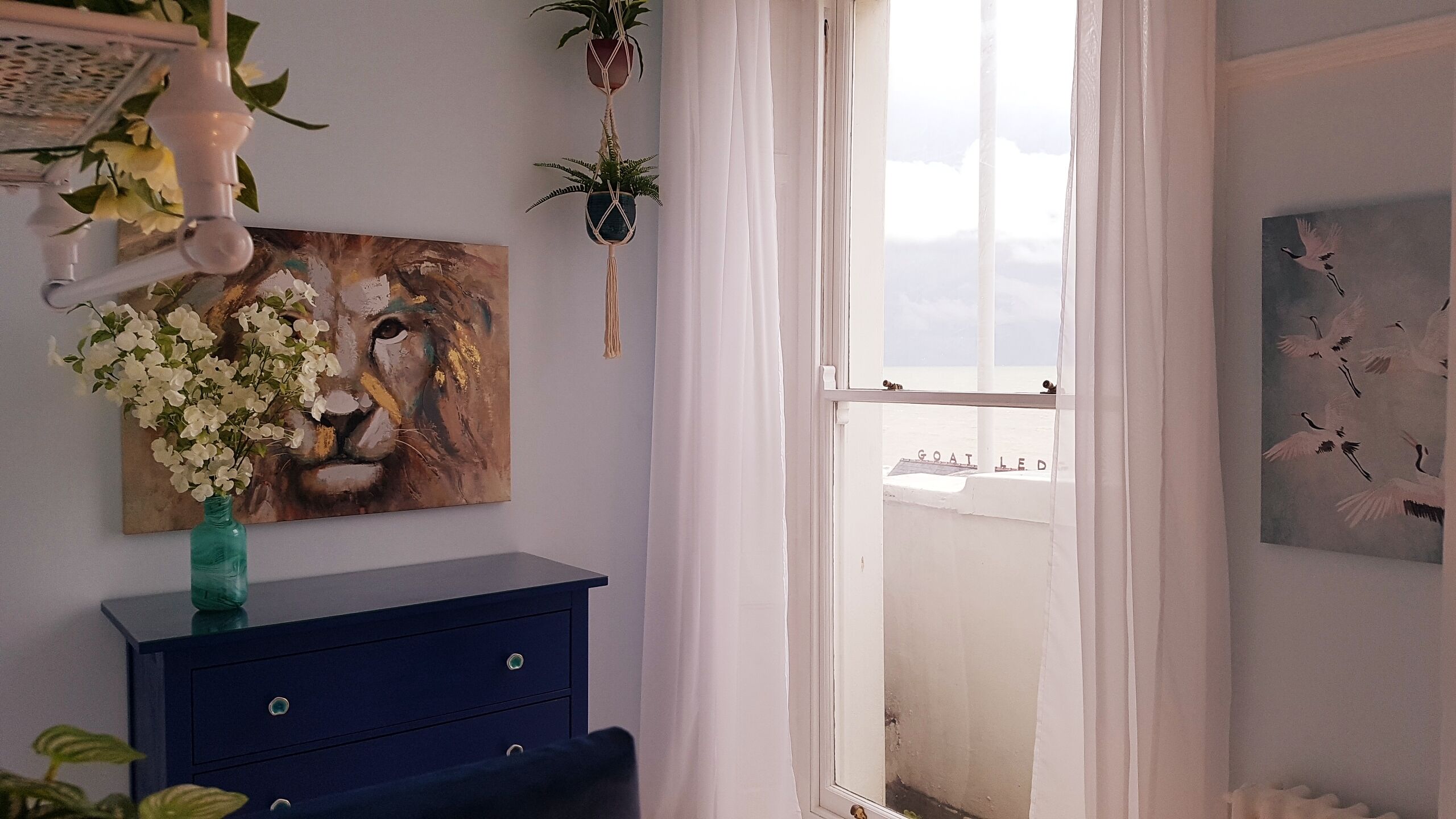 A room with a blue dresser, a painting of a lion's face, a vase with white flowers, and a view of the sea through a window.