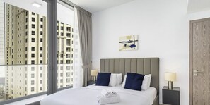 A hotel room with a large bed, two bedside tables, a window with a view of high-rise buildings, and a wooden door.