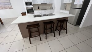 A modern kitchen with white cabinets, a stainless steel refrigerator, a built-in oven, and a sink. There are three wooden stools at the island.