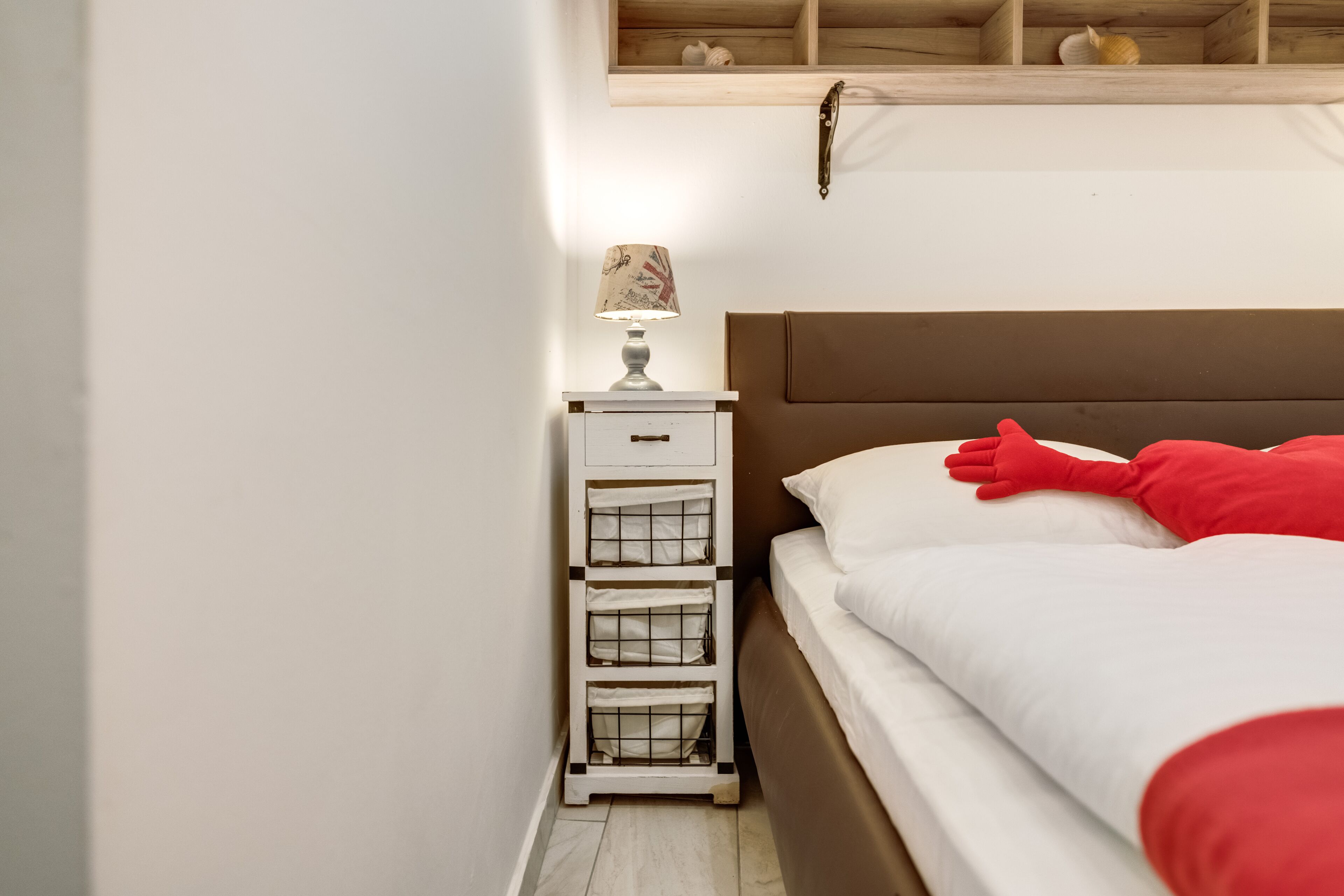 A bedroom with a bed, white nightstand with drawers, a lamp, and a wooden shelf above the bed.
