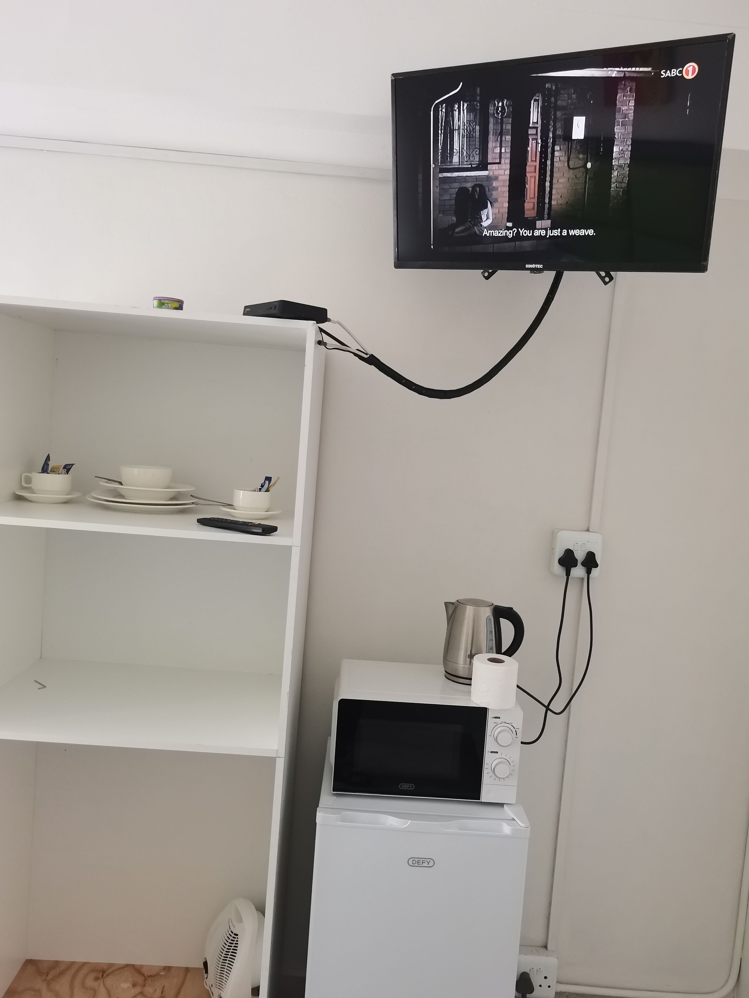 A white shelf with cups, bowls, and a remote control. A white microwave oven is on the counter.
