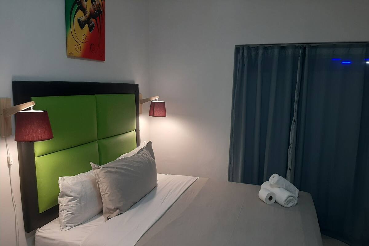 Ein Hotelzimmer mit einem grünen Stoffkopfende, weißer Bettwäsche, einer roten Lampe und einem Fenster mit Vorhängen.