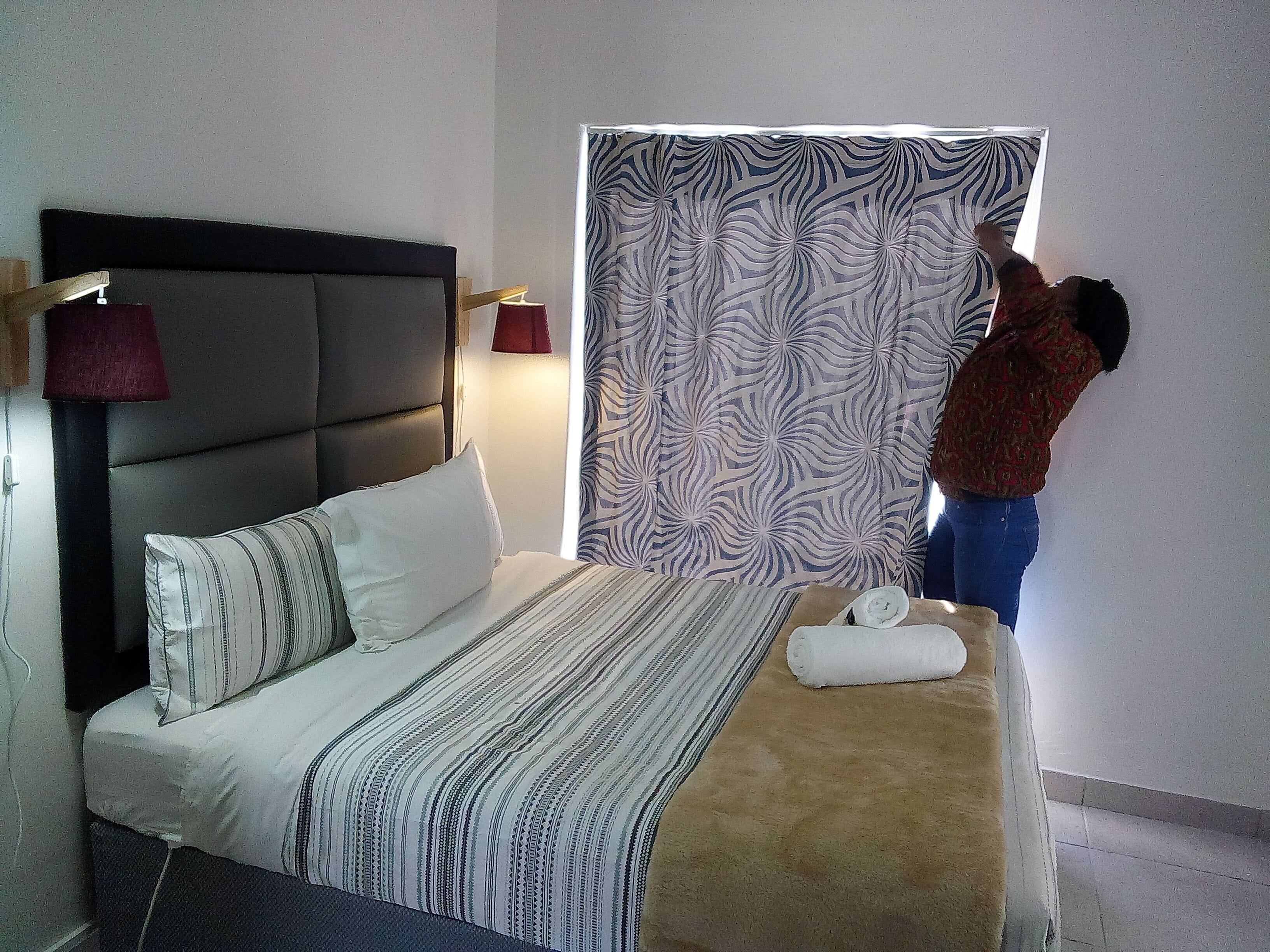 Habitación de hotel con cama, cabecera, ventana con cortinas estampadas y una toalla sobre la cama.
