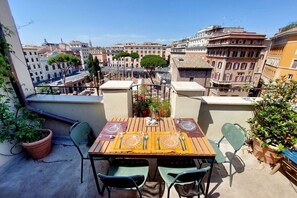 Een balkon met een houten tafel gedekt voor twee personen, omringd door stoelen, met uitzicht op een stadsbeeld met historische gebouwen.