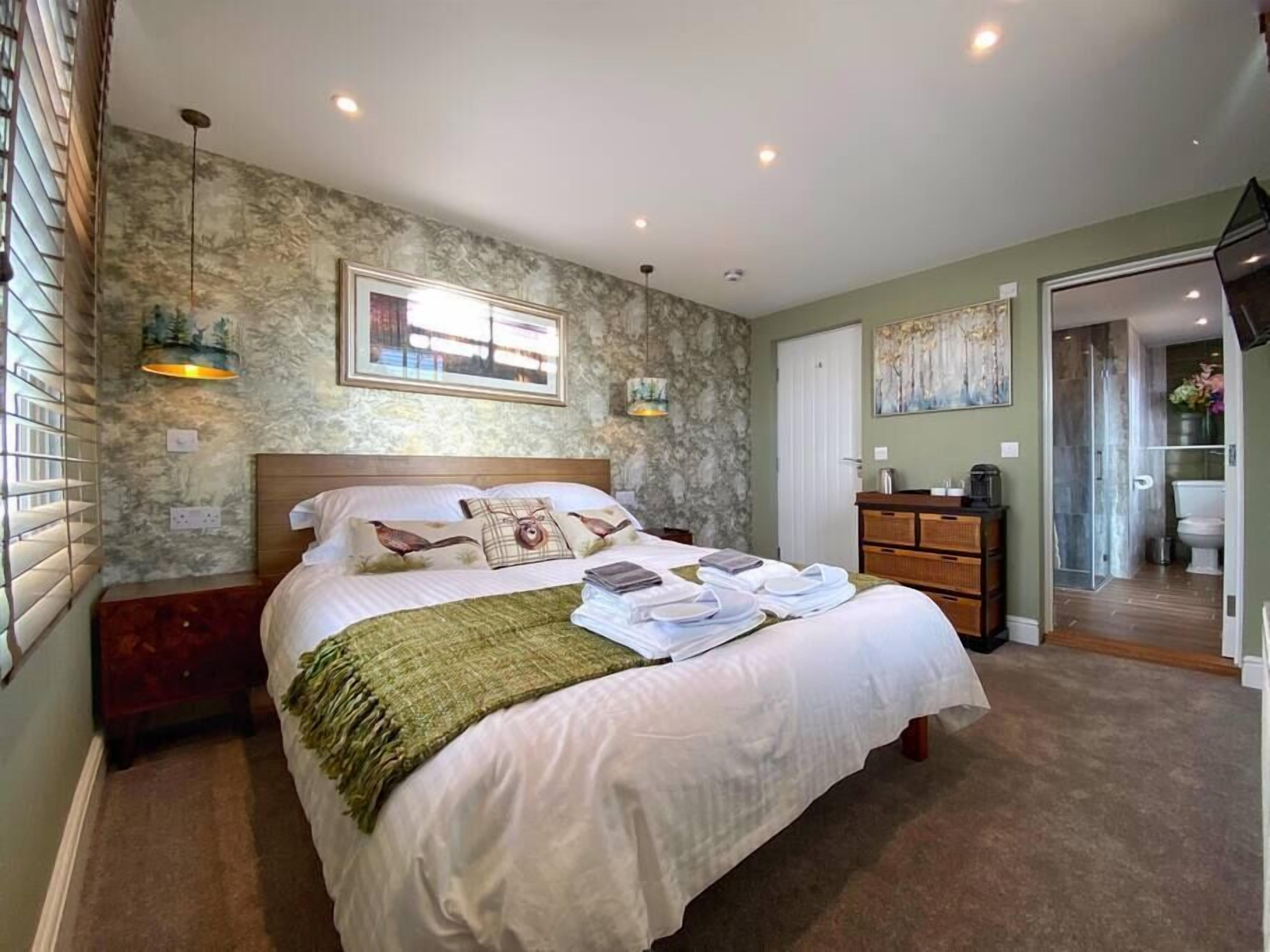 A well-lit bedroom with a large bed, a wooden nightstand, a dresser, and a bathroom visible through an open door.