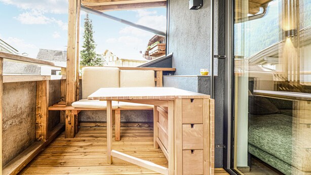 Een houten terras met een tafel, banken, een glazen deur en uitzicht op het omliggende landschap.