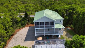 A two-story house with a green roof, surrounded by dense pine trees, and a clear view of the ocean in the background. - Heated Pool, 4 BR,  Island Blessing, Beach Chairs, Short Walk to Beach (St. George Island)