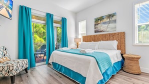 A bedroom with a large bed, teal curtains, a wicker headboard, a wicker side table, and a view of greenery through the window. - Heated Pool, 4 BR,  Island Blessing, Beach Chairs, Short Walk to Beach (St. George Island)