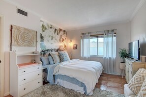 Une chambre à coucher avec un lit, une commode, un téléviseur et une fenêtre avec des rideaux.
