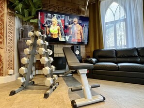 A home gym with a bench press, dumbbells, and a TV displaying fitness content. - Rare 1-bed Church Converted Apartment in London (London)