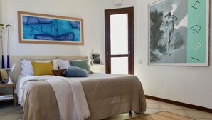 Una camera da letto con un letto, cuscini, un comodino, una lampada, un quadro astratto incorniciato e una porta.