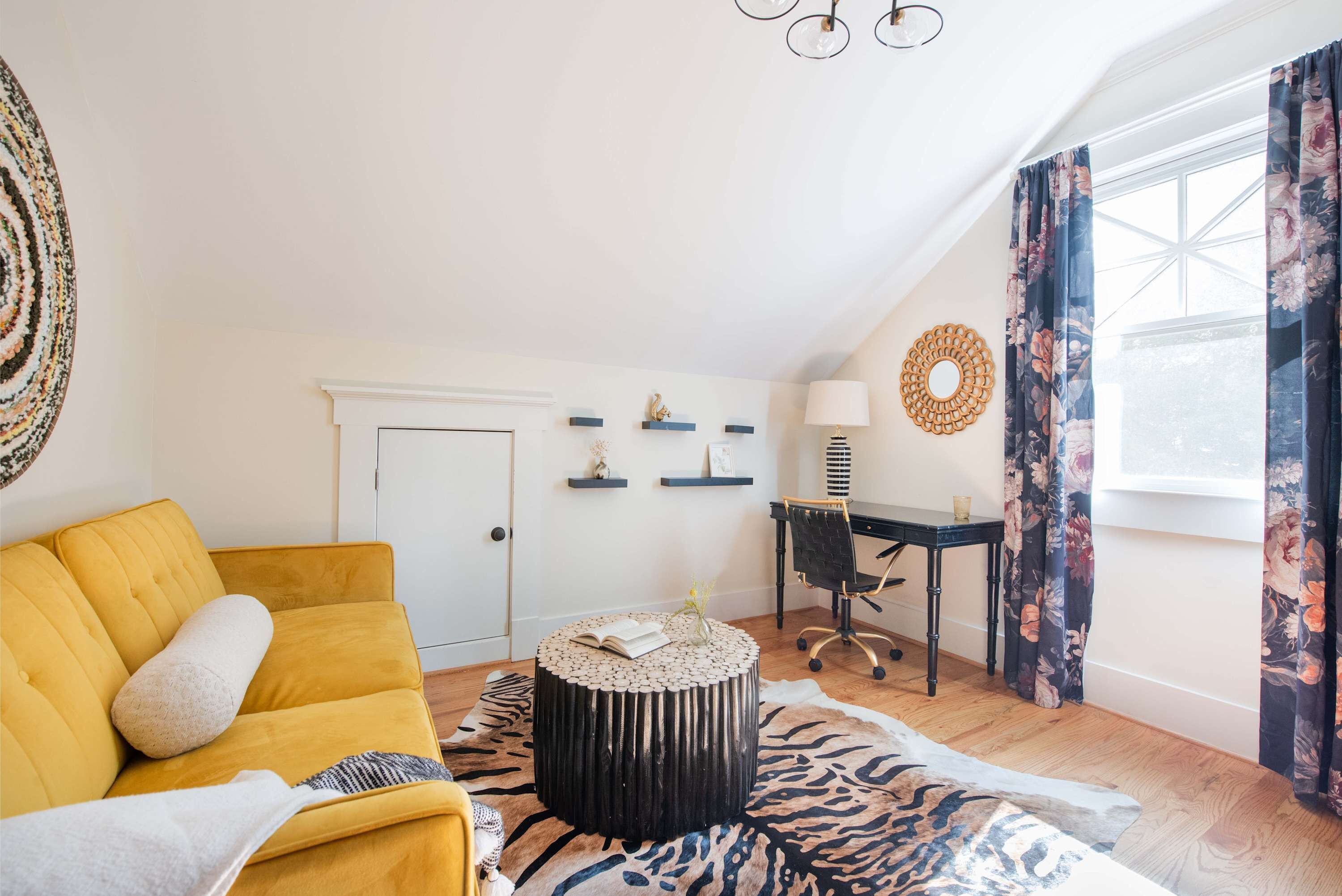 A cozy living room with a yellow sofa, a round coffee table, and a zebra-patterned rug. There is a desk with a chair, a lamp, and decorative shelves on the wall.