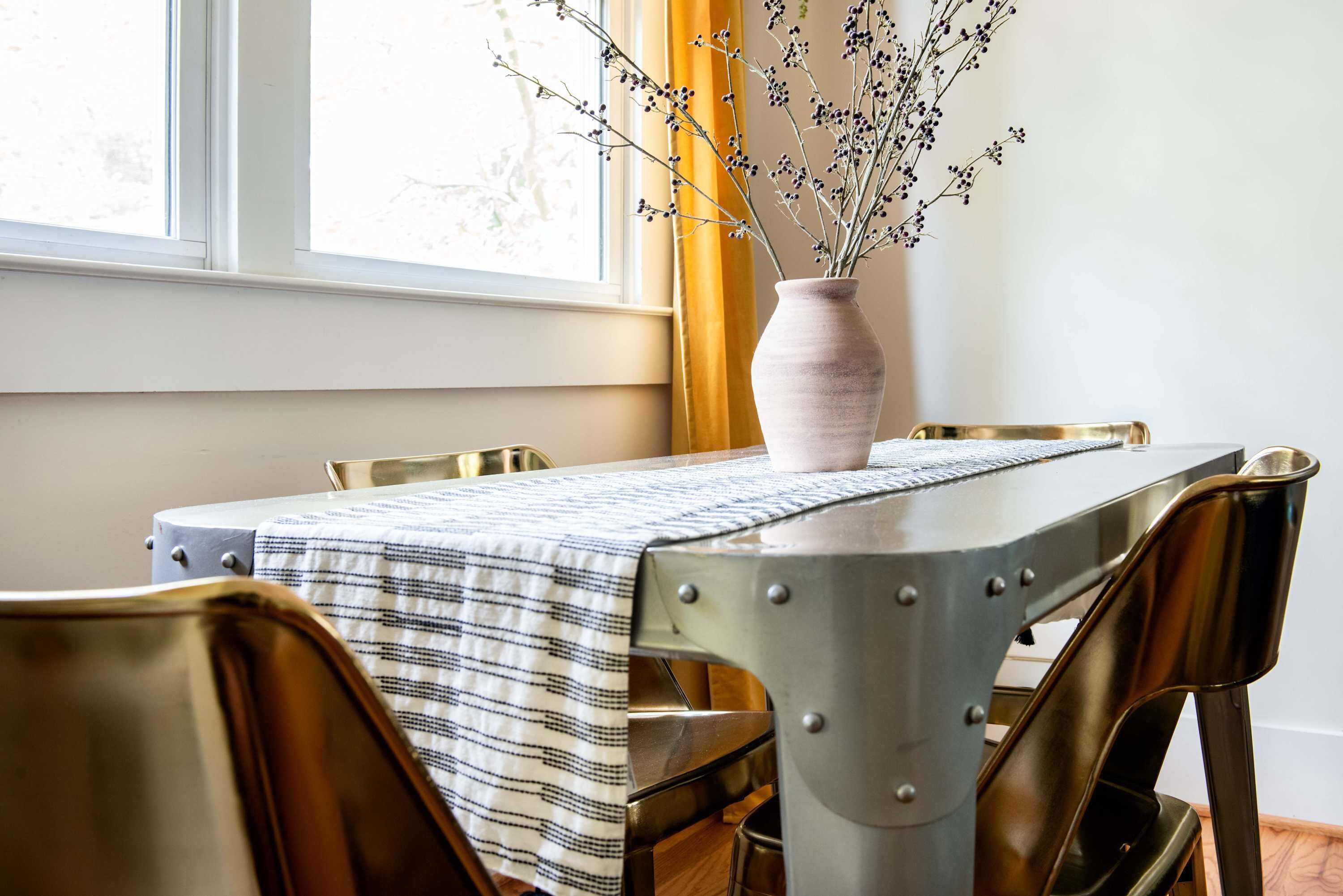 Uma mesa de jantar com toalha listrada, um vaso com galhos e uma janela com cortinas.