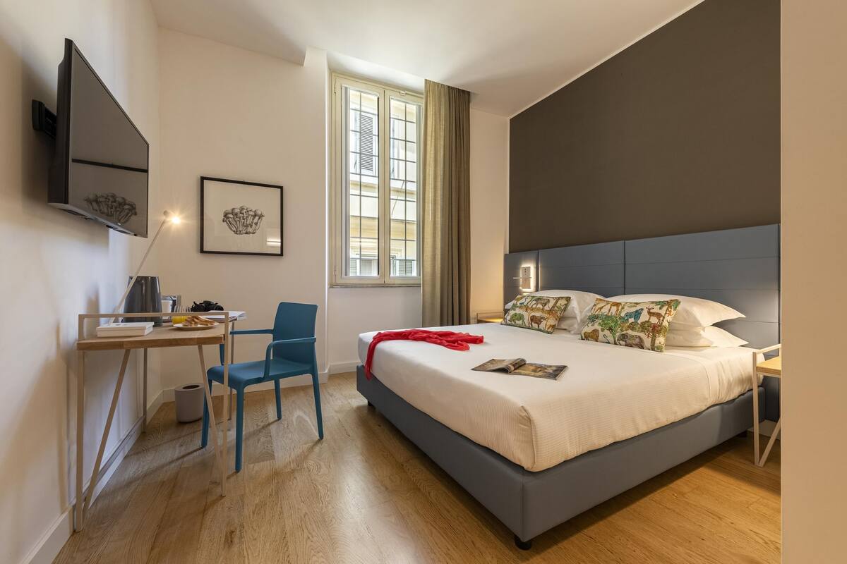 A modern bedroom with a large bed, a desk, a chair, a TV mounted on the wall, and a window with curtains.