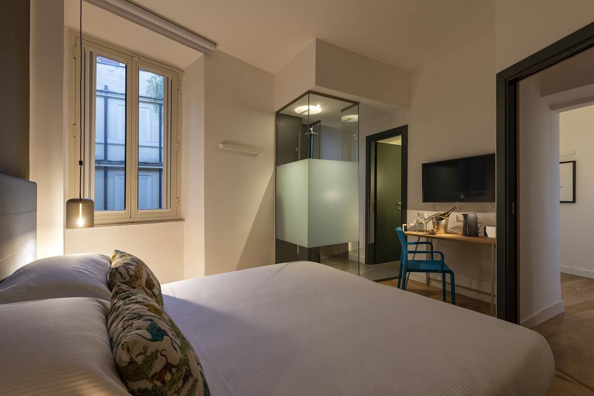 A modern hotel room with a large bed, a desk with a chair, a TV, and a bathroom with a glass shower enclosure.