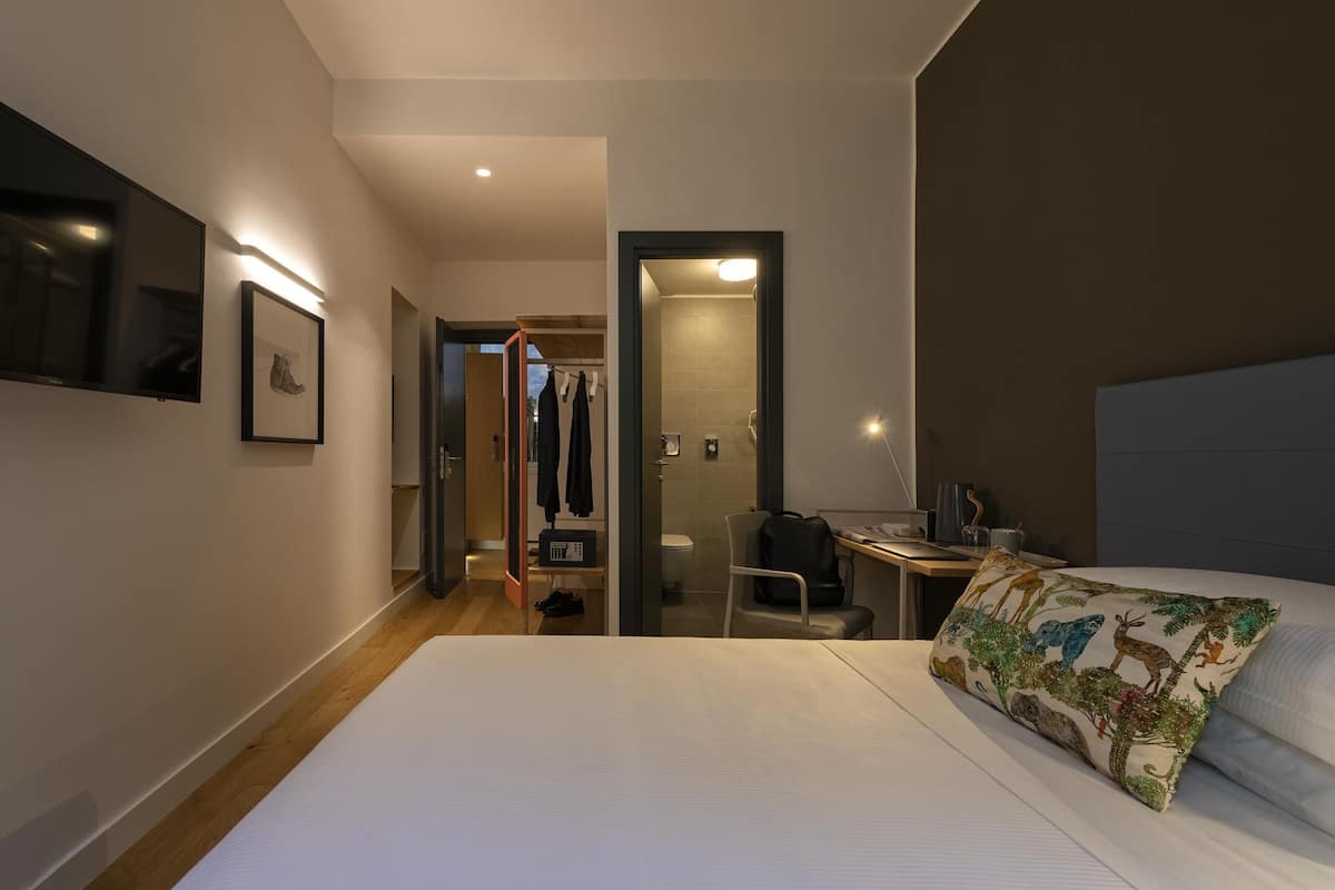 A hotel room with a large bed, a desk, and a chair. There is a closet with clothes and a shower visible through an open door.