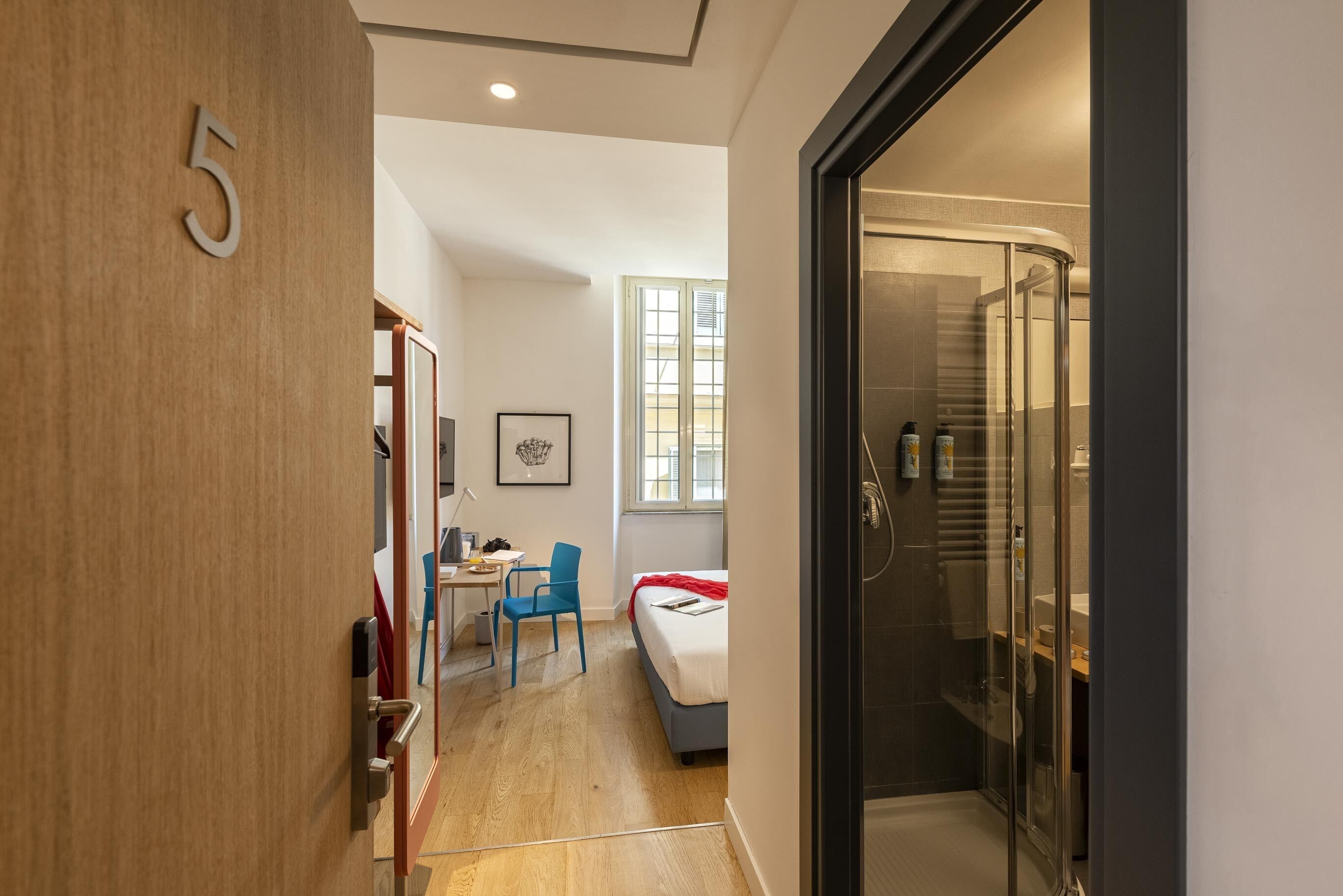 Ein modernes Hotelzimmer mit einer Holztür mit der Aufschrift '5', einer Duschkabine aus Glas, einem Bett mit roter Decke, einem Schreibtisch mit Stuhl und einem Fenster mit Jalousien.