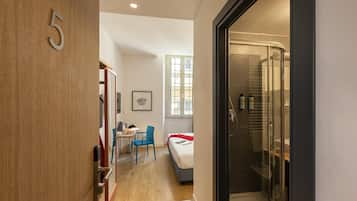 Ein modernes Hotelzimmer mit einer Holztür mit der Aufschrift '5', einer Duschkabine aus Glas, einem Bett mit roter Decke, einem Schreibtisch mit Stuhl und einem Fenster mit Jalousien.
