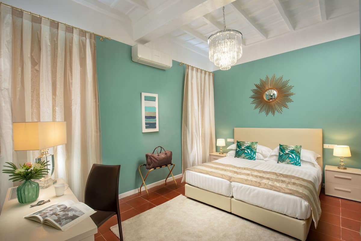 A bedroom with a bed, bedside tables, a lamp, a chair, a desk with a vase and book, and a wall-mounted mirror.