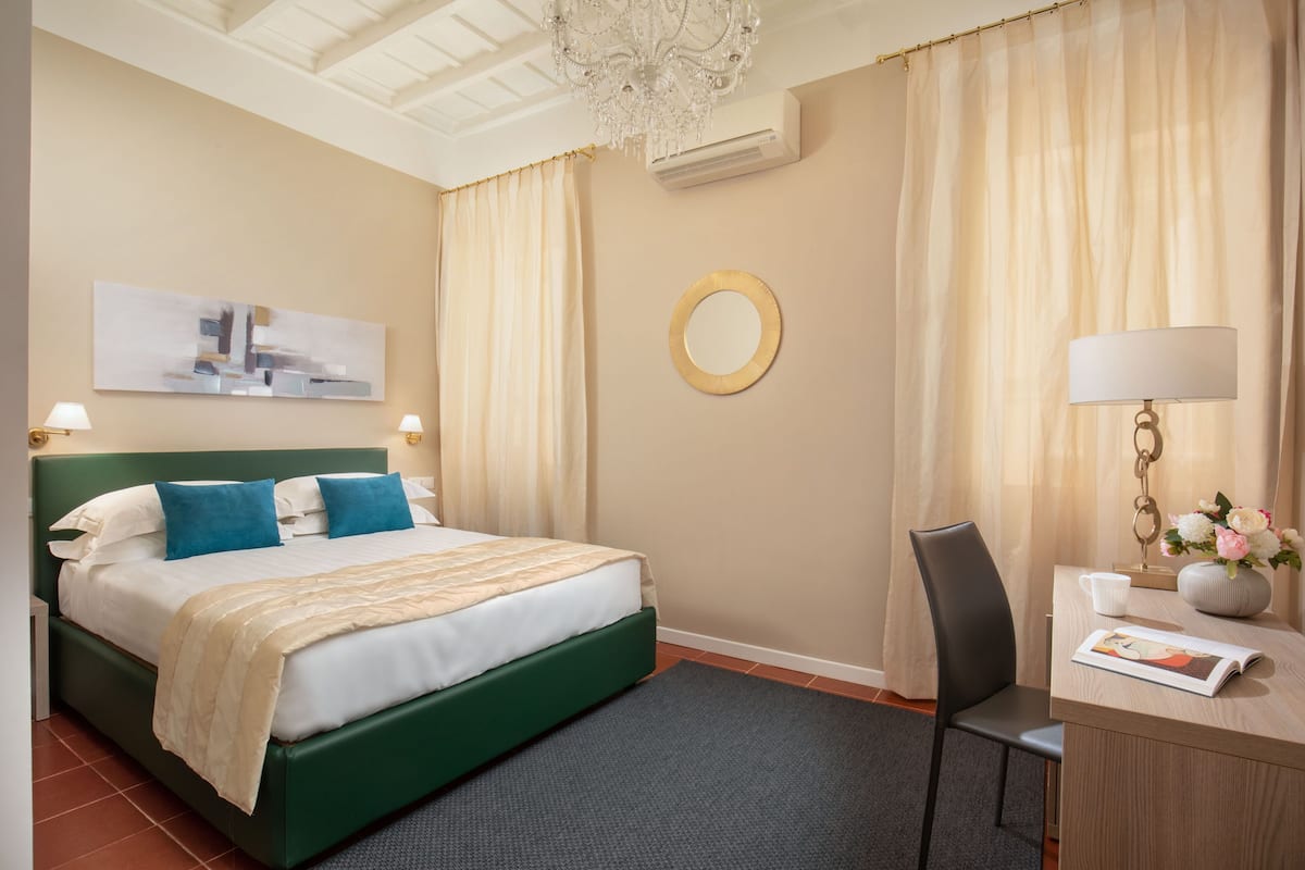 A neatly arranged hotel room with a bed, a desk with a chair, a lamp, a mirror, and a framed picture on the wall.