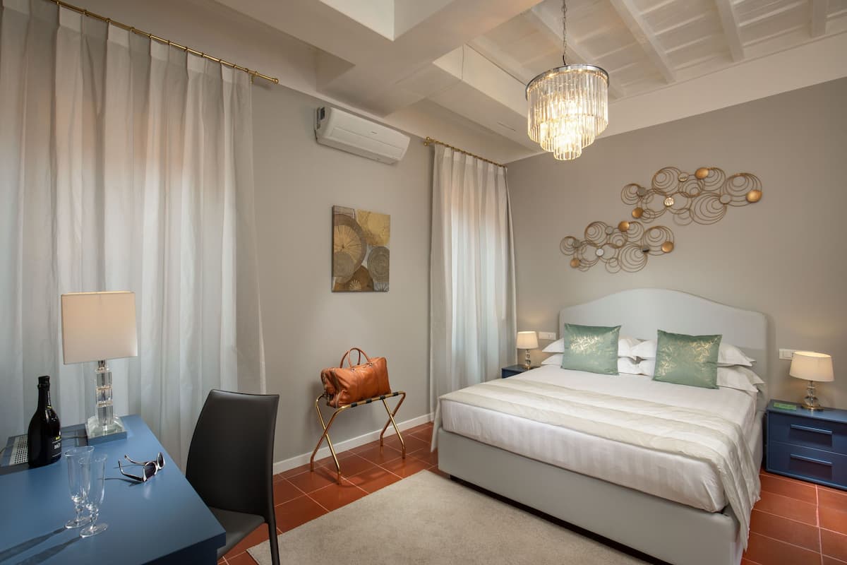 A hotel room with a bed, bedside tables, a desk with a chair, a handbag on a stand, and a chandelier.