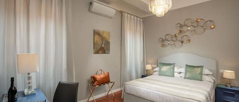 A hotel room with a bed, bedside tables, a desk with a chair, a handbag on a stand, and a chandelier.