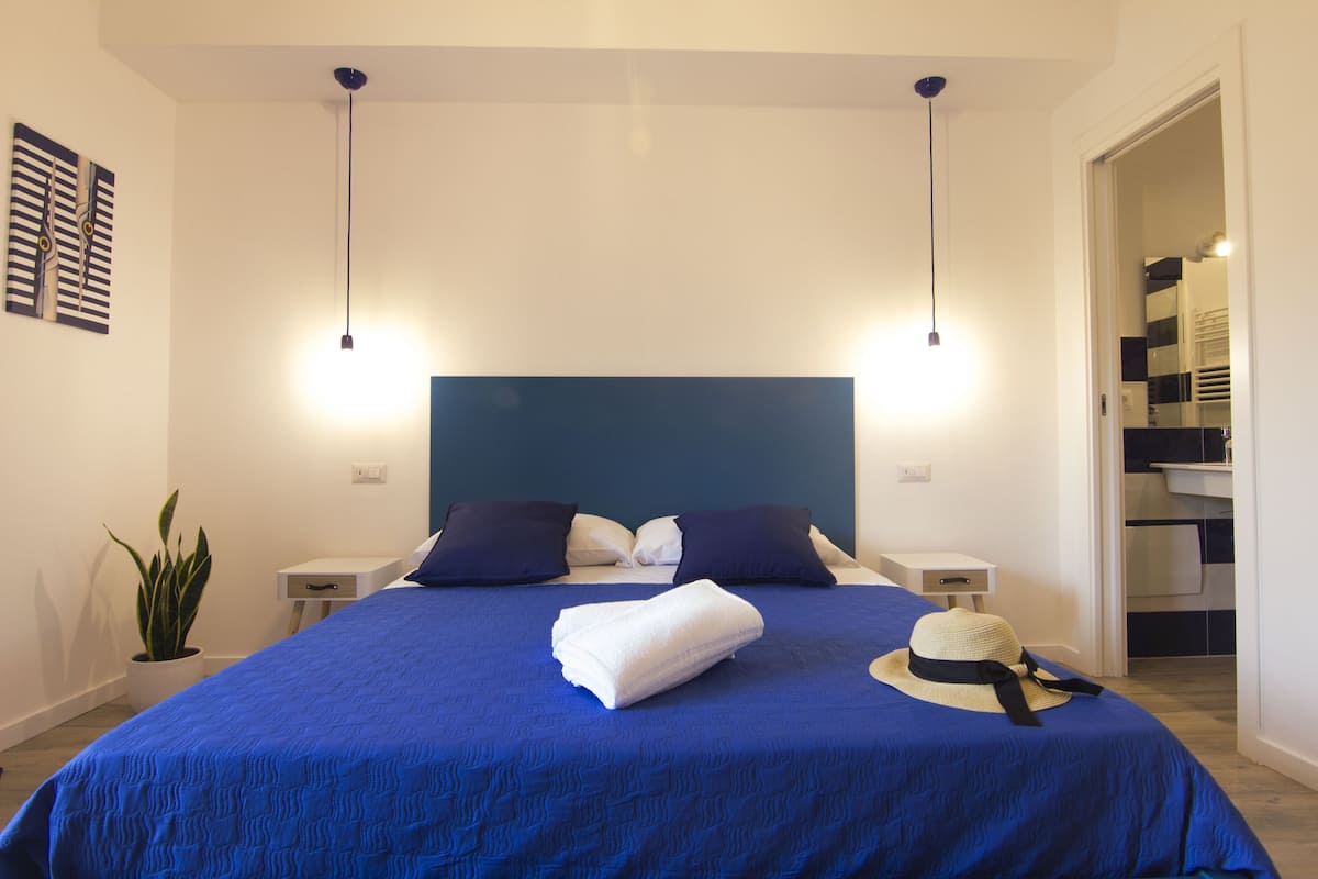 A neatly made bed with a blue bedspread, two bedside tables, a potted plant, and a wall-mounted artwork.