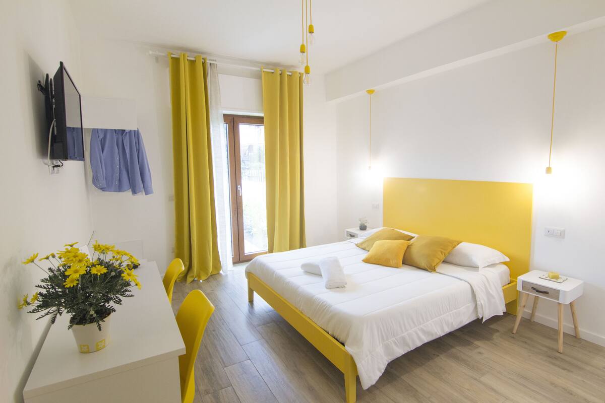A modern bedroom with a large bed, a bedside table, a chair, a desk, and a potted plant.