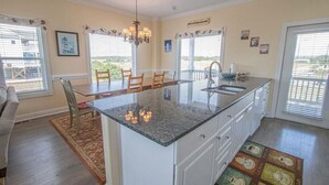 Cozinha com ilha central, armários brancos e lustre. Há cadeiras de jantar e uma mesa, e o espaço possui grandes janelas com persianas.