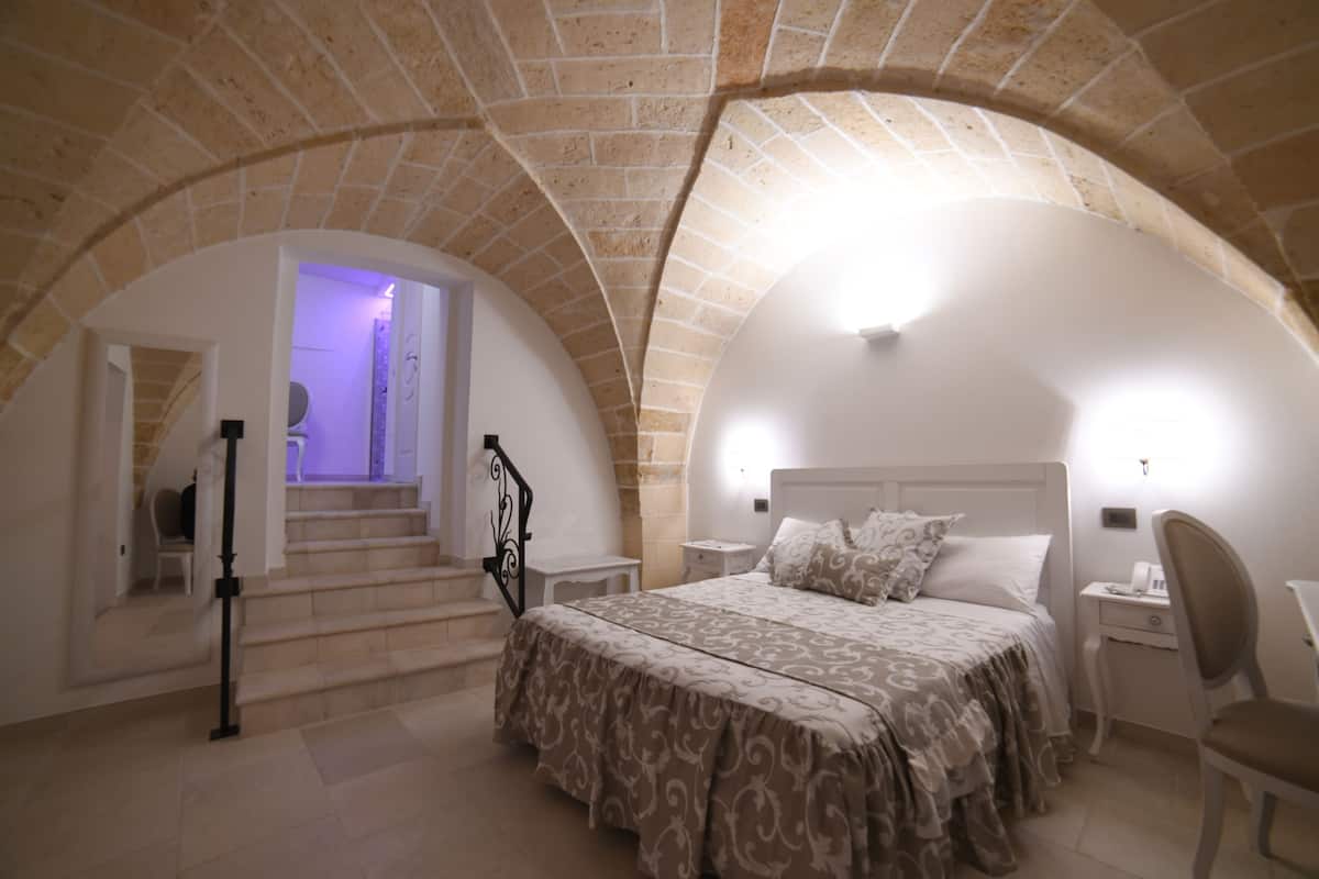 A bedroom with a bed, a desk, and a chair. There are stairs leading to an upper level. The room has a stone ceiling and walls.