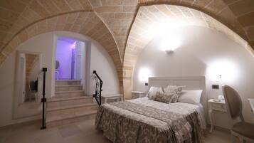 A bedroom with a bed, a desk, and a chair. There are stairs leading to an upper level. The room has a stone ceiling and walls.