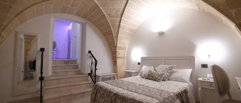 Un dormitorio con una cama, un escritorio y una silla. Se ven escaleras que suben a un nivel superior. La habitación tiene techo y paredes de piedra.