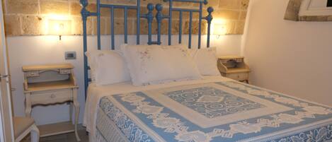 Un dormitorio con pared de piedra, una cama con colcha azul y blanca estampada, dos mesitas de noche y una mesilla.