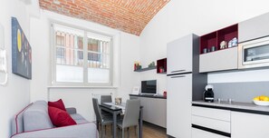 Una cocina moderna con armarios blancos, una zona de comedor con sofá gris y cojines rojos, y techo de ladrillo.