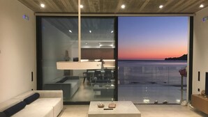 Een moderne woonkamer met een grote glazen deur die leidt naar een balkon met uitzicht op het water bij zonsondergang.