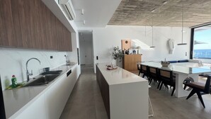 Een moderne keuken met een centraal eiland, een eetgedeelte met een glazen tafel, en uitzicht op zee.