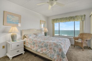 A bedroom with a bed, two bedside tables, a chair, a ceiling fan, and a window with a view of the ocean.