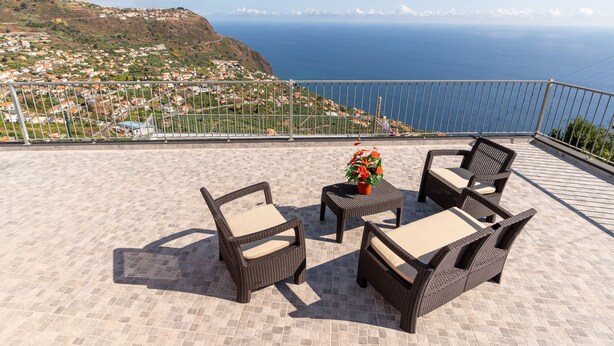 A balcony with wicker furniture and a potted plant, overlooking a coastal town and the sea.