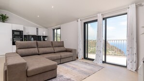 A modern living room with a sectional sofa, a kitchen in the background, and a balcony with a view.