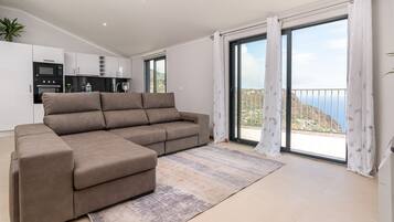 A modern living room with a sectional sofa, a kitchen in the background, and a balcony with a view.