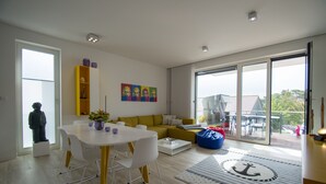 A modern living room with a yellow sofa, white dining table, and a large sliding glass door leading to a balcony.